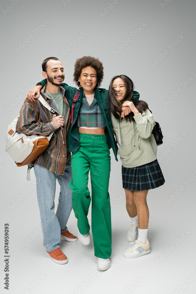 Fototapeta premium full length of joyful african american woman embracing multiethnic students with backpacks on grey background.