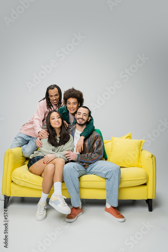 happy and stylish multicultural friends posing near yellow couch and looking at camera on grey background.