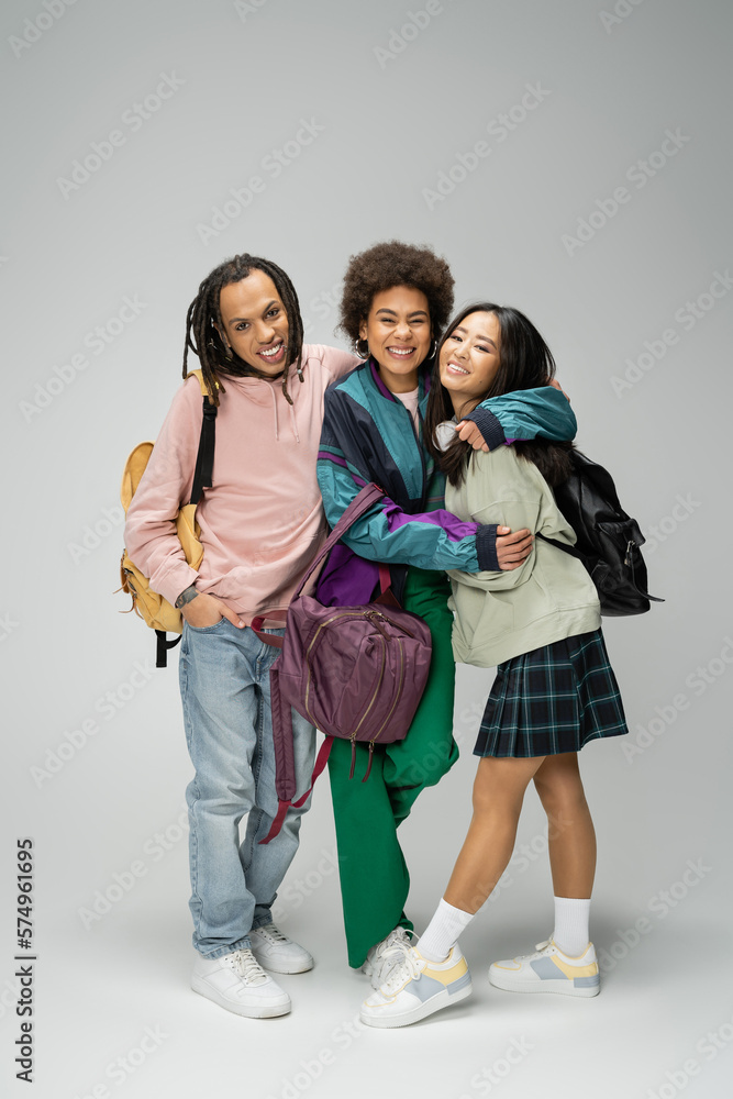 Fototapeta premium full length of joyful multicultural students with backpacks embracing while looking at camera on grey background.