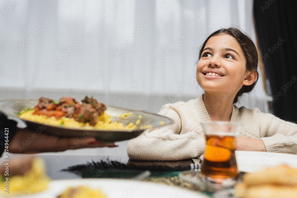 Smiling muslim girl looking at blurred mom with pilaf at home.