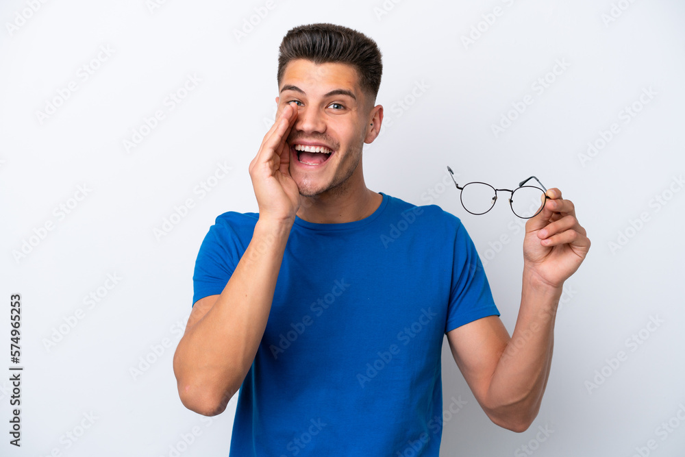 Young caucasian man holding glasses isolated on white background shouting with mouth wide open Young caucasian man holding glasses isolated on white background shouting with mouth wide open