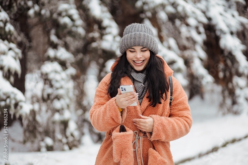 Pretty long-haired girl with smartphone at winter snow park in cold weather scrolling texting chatting using mobile phone