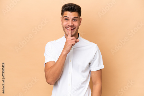 Young caucasian man isolated on beige background showing a sign of silence gesture putting finger in mouth