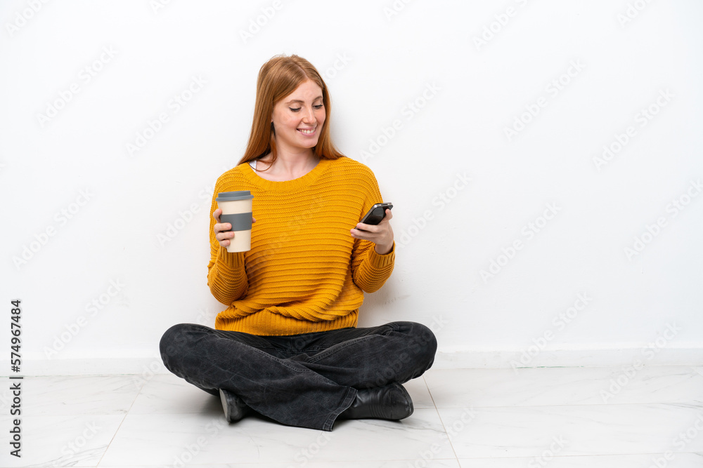 Young redhead woman sitting on the floor isolated on white background holding coffee to take away and a mobile Young redhead woman sitting on the floor isolated on white background holding coffee to take away and a mobile
