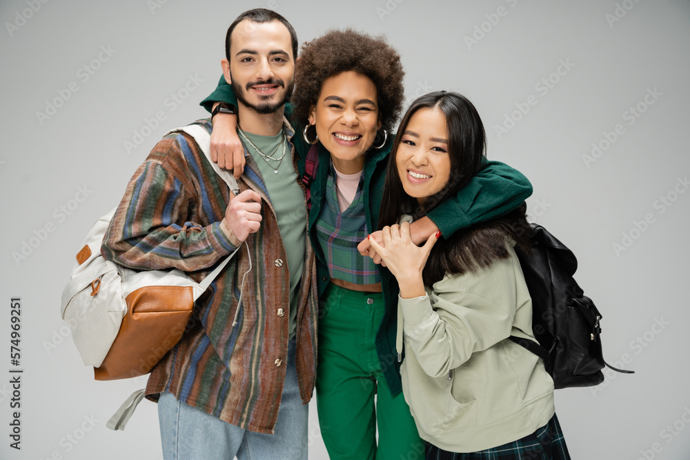 excited african american woman embracing stylish multiethnic students with backpacks isolated on grey.