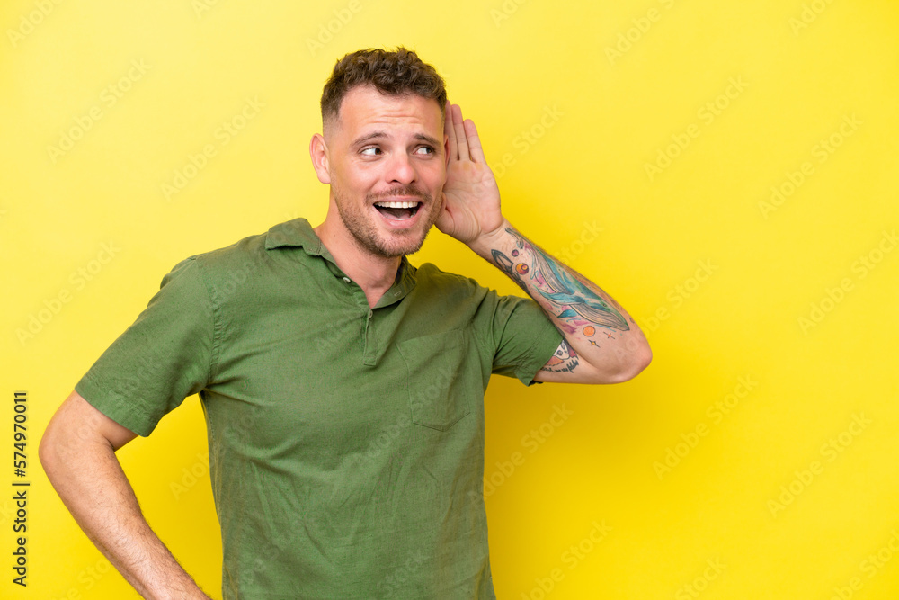Young caucasian handsome man isolated on yellow background listening to something by putting hand on the ear Young caucasian handsome man isolated on yellow background listening to something by putting hand on the ear