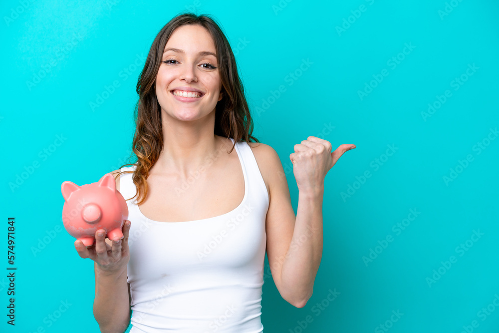 Young Caucasian woman holding a piggybank isolated on blue bakcground pointing to the side to present a product Young Caucasian woman holding a piggybank isolated on blue bakcground pointing to the side to present a product
