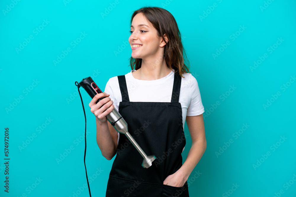 Young caucasian cooker woman using hand blender isolated on blue background looking side