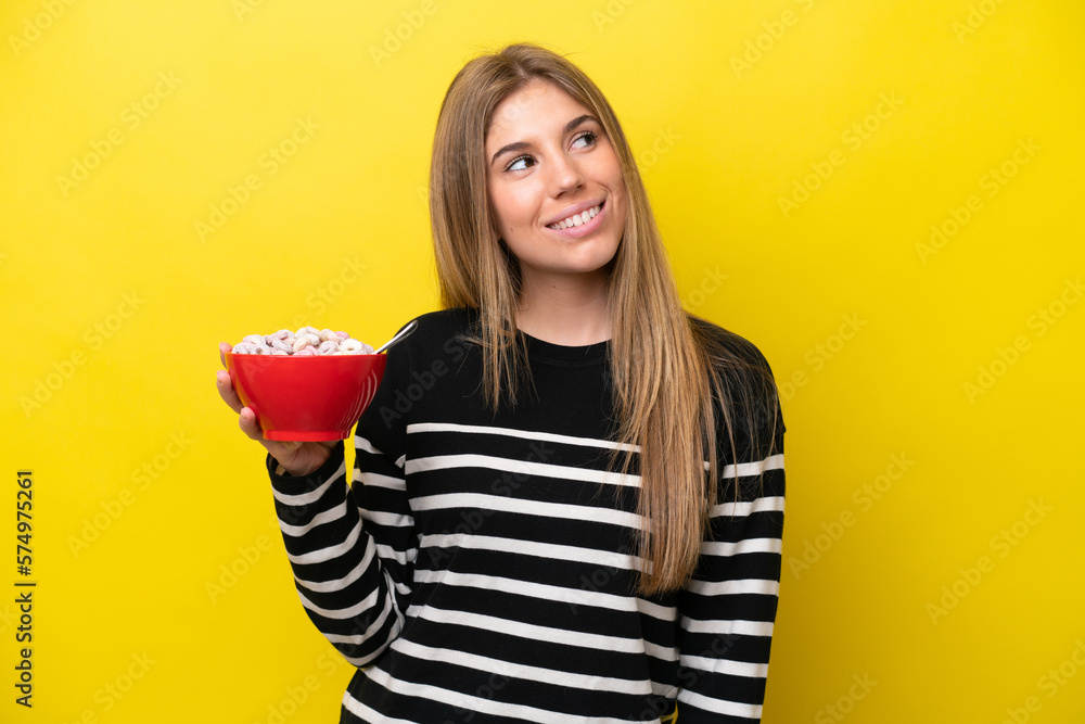 Young caucasian woman holding a bowl of cereals isolated on yellow background looking up while smiling Young caucasian woman holding a bowl of cereals isolated on yellow background looking up while smiling