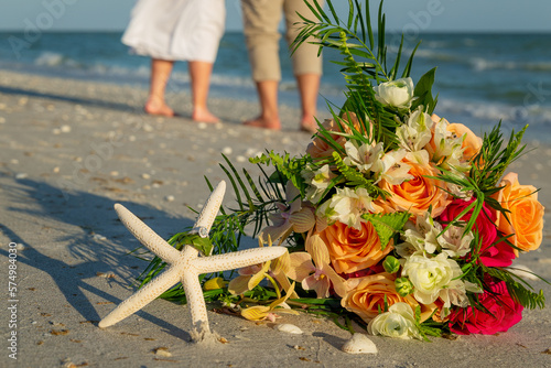 Bridal bouquet and starfish with wedding rings on sandy beach with waist down of bride and groom walking away in background photo