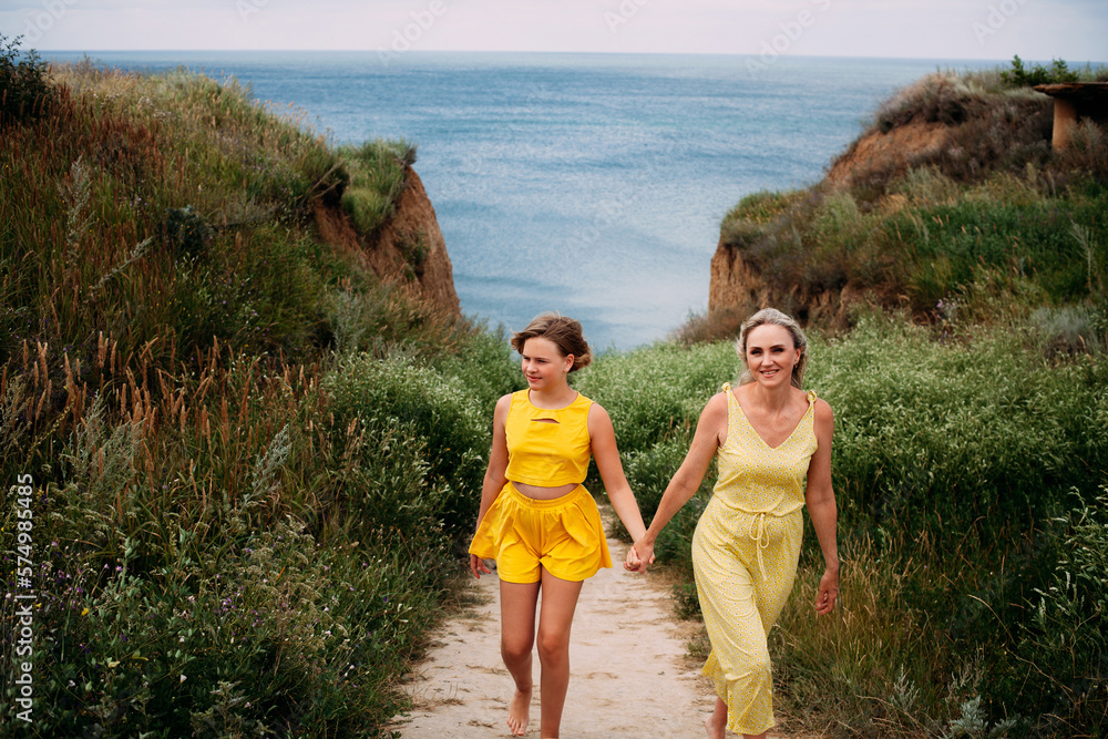 A beautiful family, a mother and daughter in yellow summer clothes walk hand in hand and smile, among the hills near the ocean on a bright sunny day. A beautiful family, a mother and daughter in yellow summer clothes walk hand in hand and smile, among the hills near the ocean on a bright sunny day.
