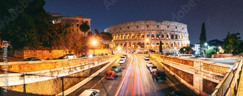 Rome, Italy. Colosseum Also Known As Flavian Amphitheatre. Traffic In Rome Near Famous World Landmark UNESCO In Evening Time. Amazing Bright Night Starry Sky Background. Dark Blue Sky. Travel Italy.