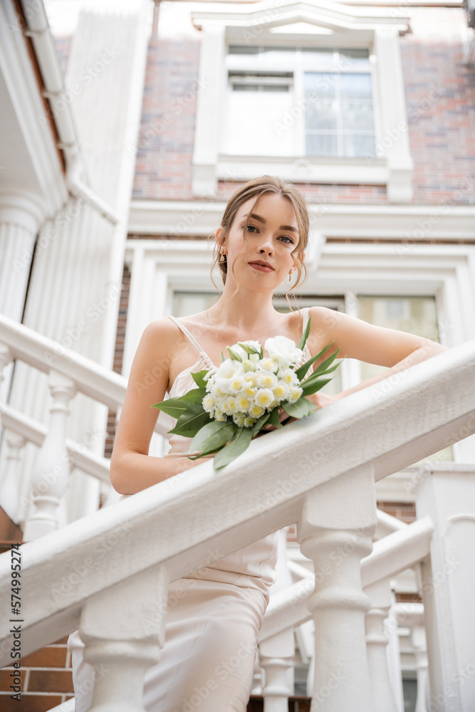 low angle view of bride in white dress holding bouquet and looking at camera near house. low angle view of bride in white dress holding bouquet and looking at camera near house.