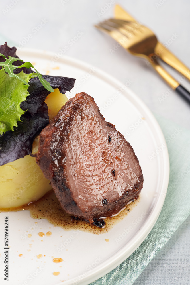 A medium rare ostrich steak in barbecue sauce with herb mashed potatoes topped with fresh green arugula leaves on a white plate on a grey background A medium rare ostrich steak in barbecue sauce with herb mashed potatoes topped with fresh green arugula leaves on a white plate on a grey background