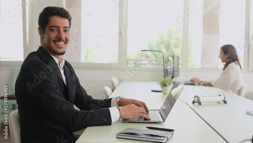 Wallpaper Mural Cheerful latin male freelancer or office worker is using laptop computer sitting in coworking space. Friendly and optimistic eastern dark-haired guy in formal suit looks at camera and smiles Torontodigital.ca