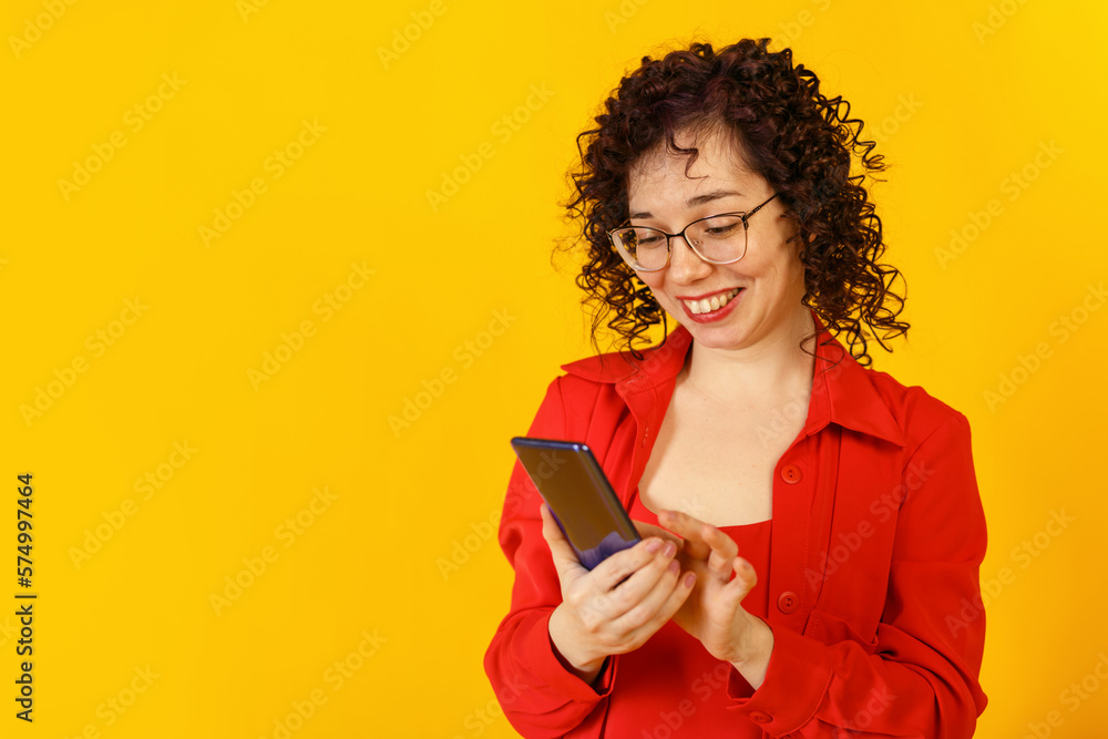 Cute curly girl with glasses is using her smartphone. Young woman in red clothes chating online and surprised. Studio female portrait on a bright yellow background. Copy space. Cute curly girl with glasses is using her smartphone. Young woman in red clothes chating online and surprised. Studio female portrait on a bright yellow background. Copy space.
