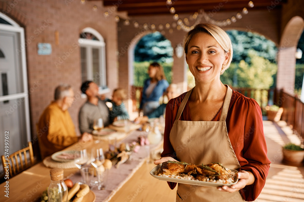 Happy woman serving food to her extended family on patio and looking at camera.