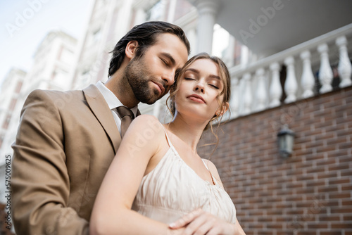 low angle view of groom in beige suit standing near gorgeous bride with closed eyes.