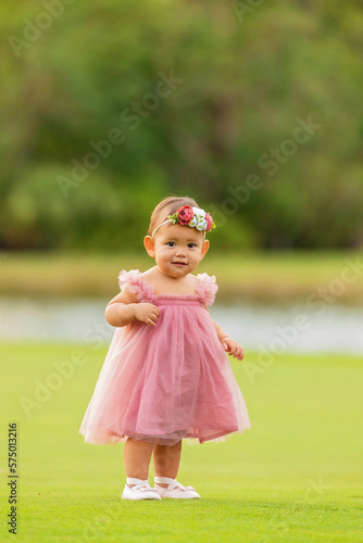 1 year old baby girl in a pink dress walking on green grass.