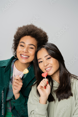 happy african american and asian women holding lollipops and looking at camera isolated on grey.
