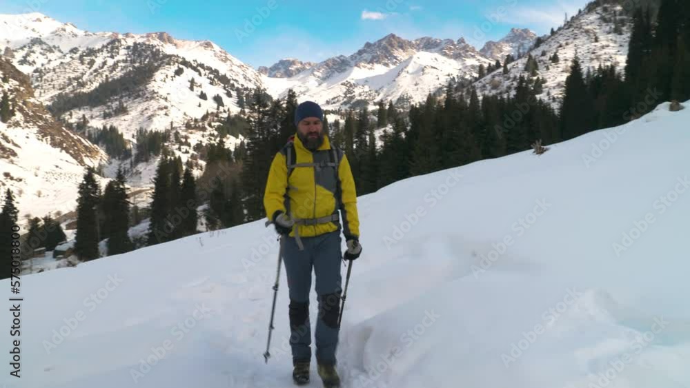 A bearded man with a backpack walks along a trail in the winter in the mountains outdoors. A tourist with a backpack and trekking poles walks along the path returning from a walk A bearded man with a backpack walks along a trail in the winter in the mountains outdoors. A tourist with a backpack and trekking poles walks along the path returning from a walk
