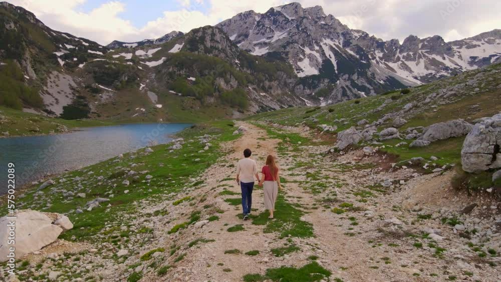 Aerial video. A young couple, dressed in casual clothes, takes a leisurely stroll through a lush valley with glacier lake and snow-capped mountains in the background. The peaceful surroundings set the Aerial video. A young couple, dressed in casual clothes, takes a leisurely stroll through a lush valley with glacier lake and snow-capped mountains in the background. The peaceful surroundings set the