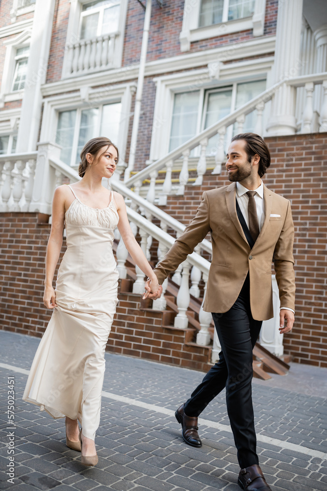 full length of happy newlyweds smiling and holding hands while walking on street.
