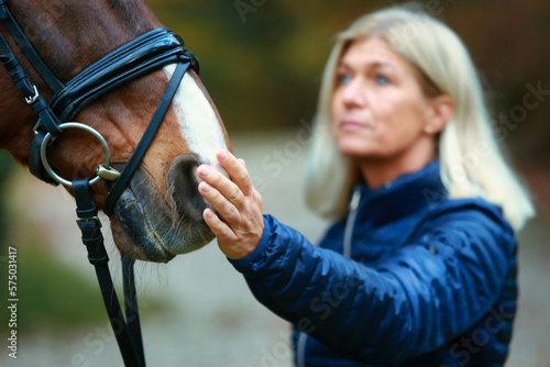 Horse head with bridle, close-up of the horse's mouth, out of focus in the background the rider, she tenderly lays her hand on the horse's mouth.. © RD-Fotografie