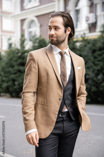young groom in beige formal wear standing with hand in pocket and looking away on street.