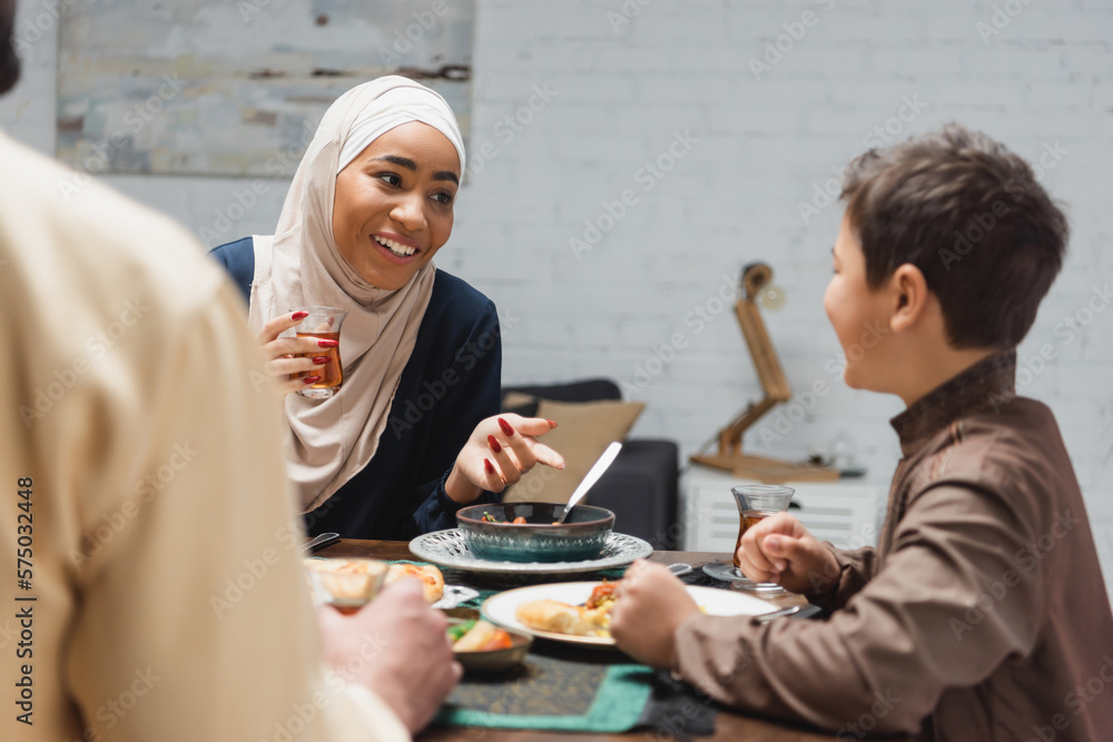 African american woman in hijab holding tea and talking to son near husband and food at home. African american woman in hijab holding tea and talking to son near husband and food at home.