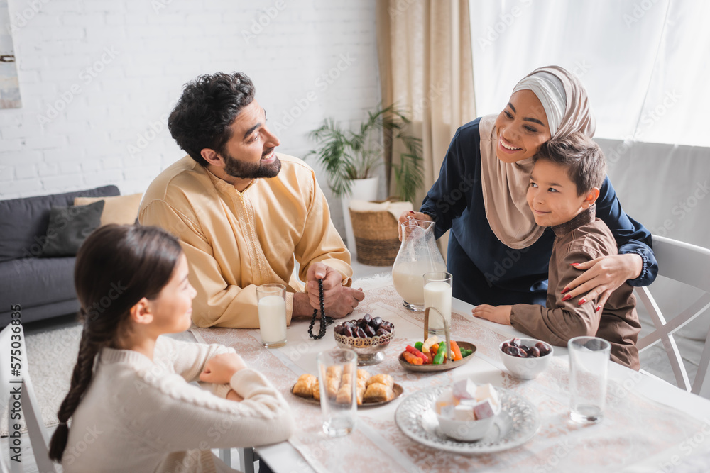 Smiling african american woman in hijab hugging son near family during suhur breakfast in morning.