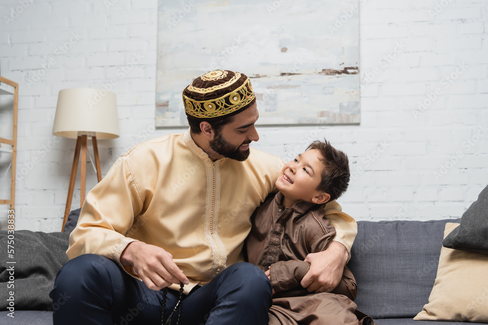Muslim man with prayer beads hugging son on couch at home.