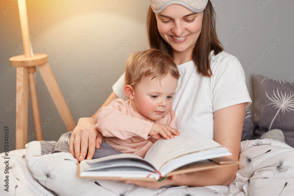 Indoor shot of smiling european woman wearing white t-shirt and blindfold sitting in bed with her toddler daughter, mommy reading fairy tale for kid, holding book in hands. Indoor shot of smiling european woman wearing white t-shirt and blindfold sitting in bed with her toddler daughter, mommy reading fairy tale for kid, holding book in hands.