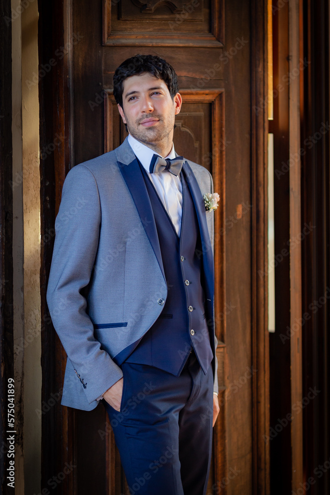 Portrait of an elegant groom leaning against an antique wooden door before going to the altar.