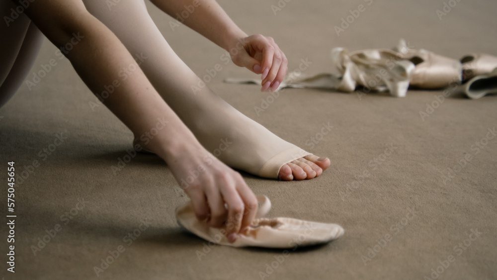 Beautiful ballet dancer sits on floor near pointe at dance studio and puts on ballet shoes before choreography lesson. Ballerina prepares for performance. Classical ballet dance school. Feet close up.