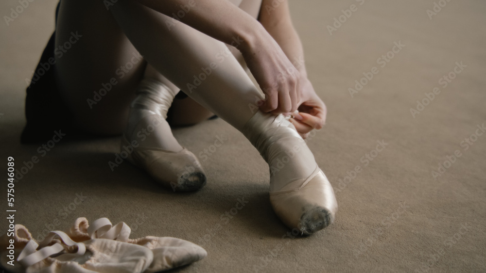 Adult ballerina sits on floor at dance studio and puts on pointe shoes before choreography lesson. Beautiful dancer prepares for performance or training. Classical ballet dance school. Feet close up. Adult ballerina sits on floor at dance studio and puts on pointe shoes before choreography lesson. Beautiful dancer prepares for performance or training. Classical ballet dance school. Feet close up.
