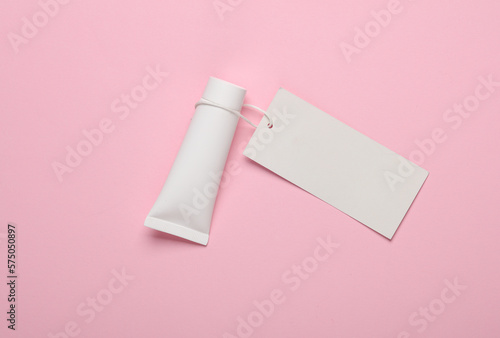 White cream tube with an empty price tag on a string, pink background. Beauty layout