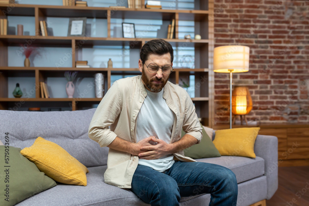 A young man feels severe stomach pain, spasms, poisoning, discomfort. Sitting on the sofa at home and holding his stomach.