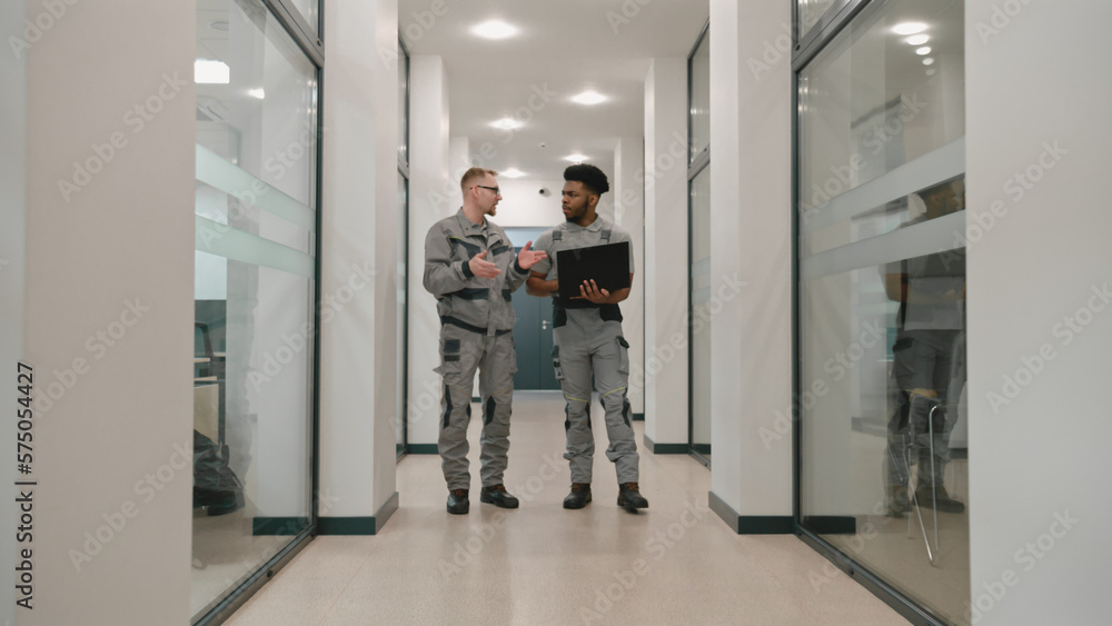 Two diverse men in uniform walk corridor and discuss CCTV cameras installation in coworking office. Installers set up security cameras using computer program on laptop. Concept of surveillance system. Two diverse men in uniform walk corridor and discuss CCTV cameras installation in coworking office. Installers set up security cameras using computer program on laptop. Concept of surveillance system.