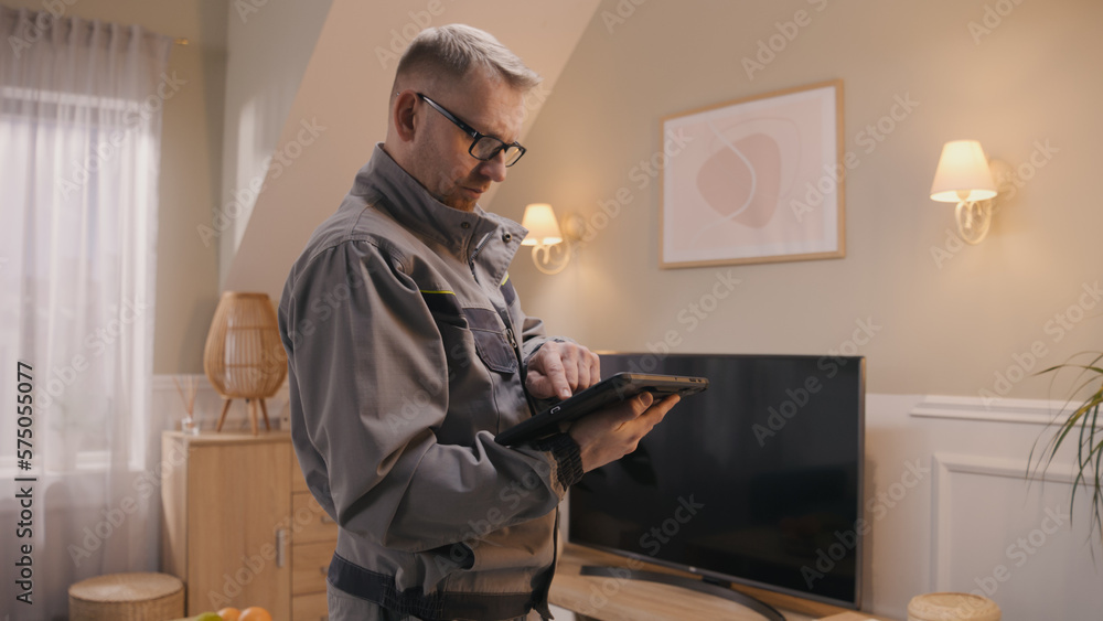 Male installer in uniform sets up security camera in apartment using digital tablet computer. Man checks CCTV camera in application. Concept of tracking and monitoring system, safety and privacy.