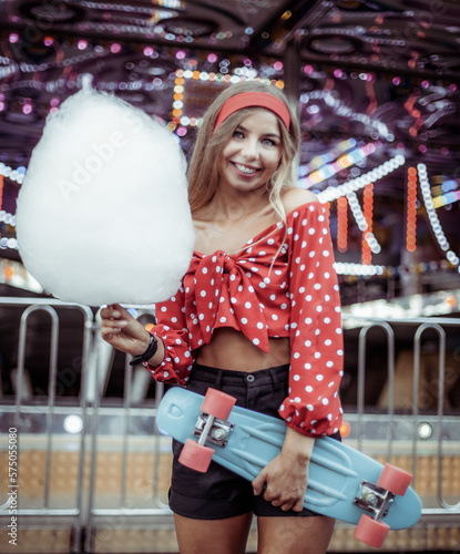 Adorable cute beautiful woman with cotton candy and penny bord stands in the middle of amusement park with bright colors, positive and cheerful, happy and optimistic emotion