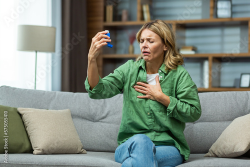 An elderly beautiful woman suffers from asthma and allergies. She is sitting sick at home on the sofa, holding an inhaler in her hands, holding her chest with her hand, suffocating, feeling pain.