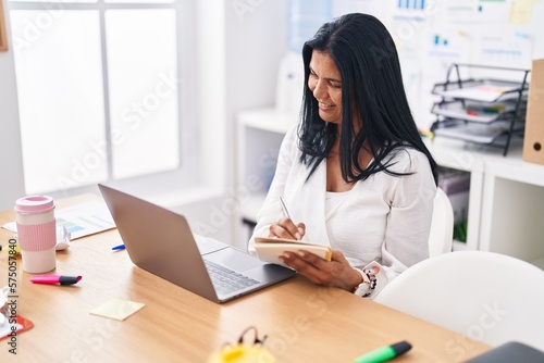 Middle age hispanic woman business worker using laptop writing on notebook at office