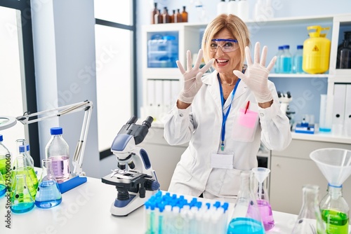 Middle age blonde woman working at scientist laboratory showing and pointing up with fingers number nine while smiling confident and happy.