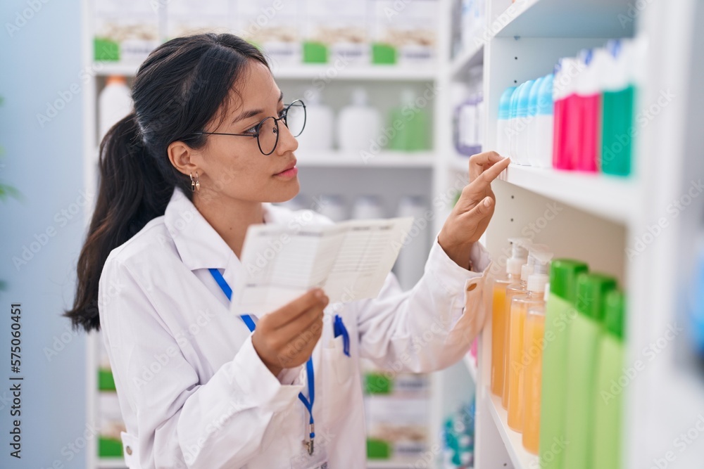 Young beautiful hispanic woman pharmacist reading prescription looking shelving at pharmacy