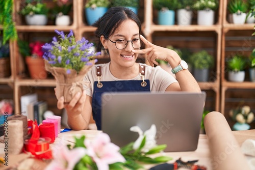 Wallpaper Mural Young hispanic woman working at florist shop doing video call doing peace symbol with fingers over face, smiling cheerful showing victory Torontodigital.ca