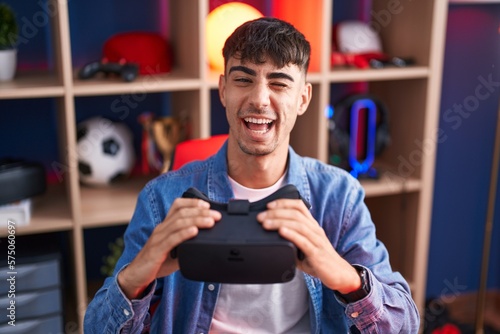 Young hispanic man wearing virtual reality glasses winking looking at the camera with sexy expression, cheerful and happy face.