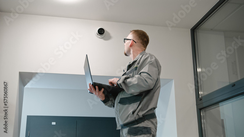 Installer in uniform sets up security camera in office room using laptop. Man in glasses checks CCTV cameras in computer program. Monitoring system. Concept of surveillance system and privacy.