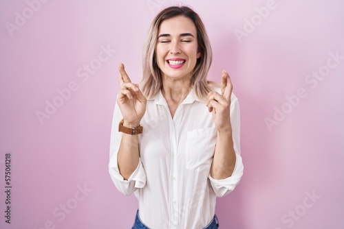 Young beautiful woman standing over pink background gesturing finger crossed smiling with hope and eyes closed. luck and superstitious concept.