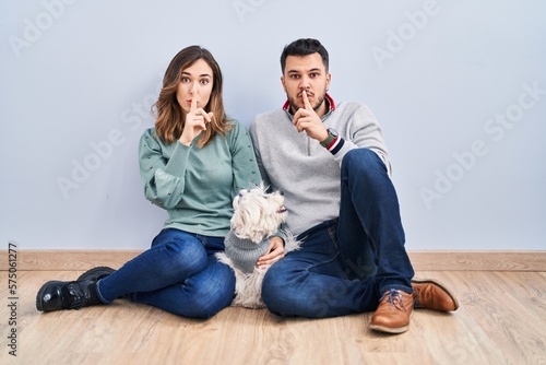 Young hispanic couple sitting on the floor with dog asking to be quiet with finger on lips. silence and secret concept.